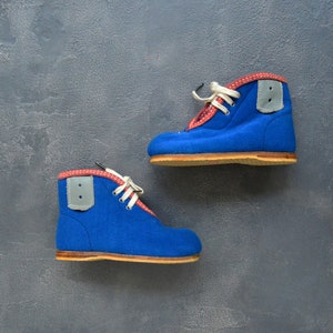May include: A pair of vintage blue baby boots with red trim and white laces. The boots have a gray patch on the side and a wooden sole. The boots are displayed against a gray background.