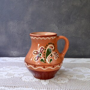 Könnte beinhalten: Handgefertigter Krug aus Terrakotta-Keramik mit Henkel. Verziert mit floralen und Blattmotiven in Weiß, Grün und Braun. Der Boden ist dunkelbraun, der Rand mit einem weißen Wellenmuster. Ein dekoratives Element.