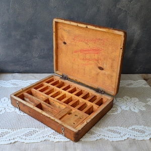 May include: A rectangular wooden storage box with a hinged lid, featuring an interior divided into multiple small compartments and a larger open section. The inside of the lid displays faint, illegible handwritten text in red ink.