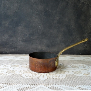 May include: A small, copper saucepan with a long, brass handle. The pot has a dark interior and a slightly aged appearance. The handle is attached with a brass rivet. The saucepan is sitting on a white lace tablecloth.