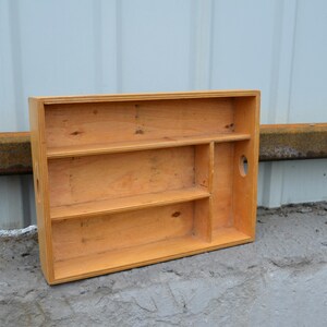 Op de afbeelding: Een rechthoekige houten plank met drie planken en een verticale verdeler. De plank is gemaakt van lichtgekleurd hout en heeft een handgreepvormige uitsparing aan de zijkant. De plank staat op een betonnen oppervlak.