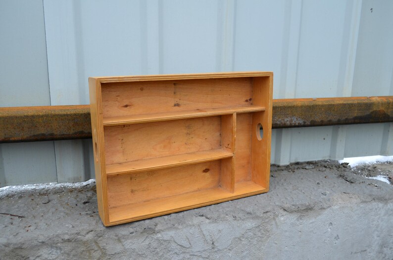 Op de afbeelding: Een rechthoekige houten plank met drie planken en een verticale verdeler. De plank is gemaakt van lichtgekleurd hout en heeft een handgreepvormige uitsparing aan de zijkant. De plank staat op een betonnen oppervlak.