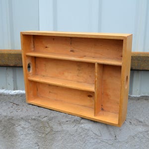 Op de afbeelding: Een lichtgekleurd houten wandrek met twee planken en een verticale verdeler. Het rek heeft een rechthoekige vorm met een gat ter grootte van een handvat aan de zijkant. Het hout heeft een natuurlijke afwerking en het rek staat op een betonnen oppervlak.