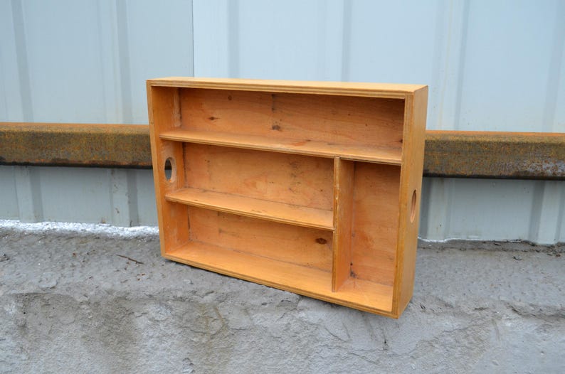 Op de afbeelding: Een lichtgekleurd houten wandrek met twee planken en een verticale verdeler. Het rek heeft een rechthoekige vorm met een gat ter grootte van een handvat aan de zijkant. Het hout heeft een natuurlijke afwerking en het rek staat op een betonnen oppervlak.