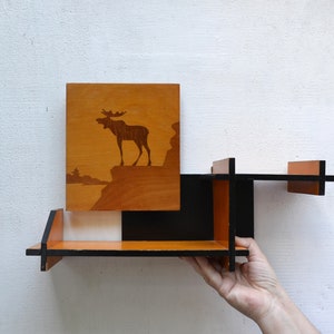 Peut inclure: Une étagère murale en bois dotée d'un panneau arrière rectangulaire orné de la silhouette brun foncé d'un élan sur une falaise. La structure de l'étagère se compose de planches en bois orange et noir entrecroisées formant plusieurs niveaux.