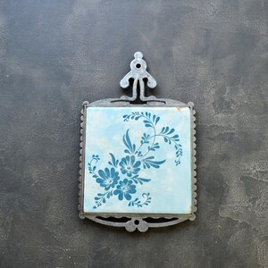 Puede incluir: Un salvamanteles cuadrado y vintage con un diseño floral azul claro sobre una baldosa de cerámica. La baldosa está enmarcada en un marco de metal ornamentado con un asa. Un elemento decorativo para la cocina.