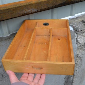 Op de afbeelding: Een rechthoekige houten lade met meerdere compartimenten en een handvat. De lade is gemaakt van lichtgekleurd hout en heeft een rond gat in de bovenkant. De lade wordt door een hand vastgehouden.