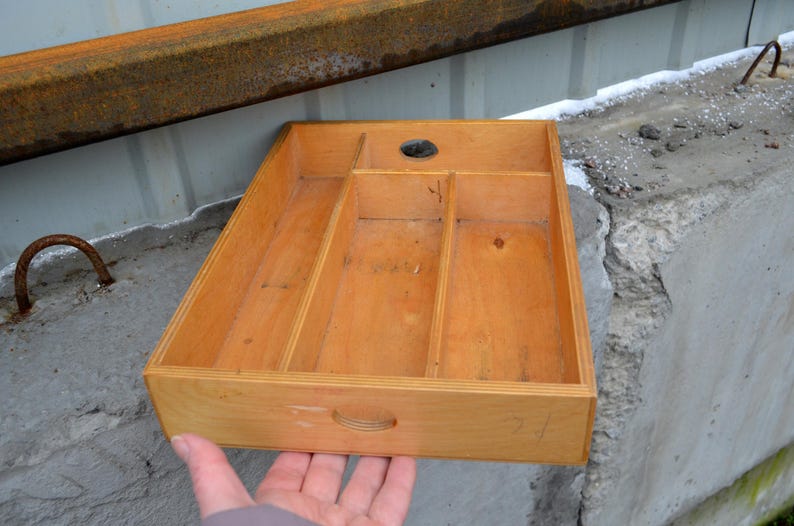 Op de afbeelding: Een rechthoekige houten lade met meerdere compartimenten en een handvat. De lade is gemaakt van lichtgekleurd hout en heeft een rond gat in de bovenkant. De lade wordt door een hand vastgehouden.