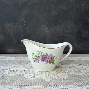 Peut inclure: Une saucière en céramique blanche ornée d'un motif floral délicat représentant des fleurs de lilas violettes et des feuilles vertes sur le côté. Le bord du récipient est souligné d'un fin liseré doré, avec une anse courbée et un bec verseur.