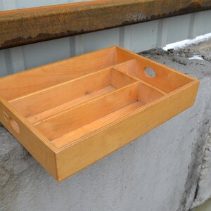 Op de afbeelding: Een lichtgekleurde houten dienblad met drie interne compartimenten en een cirkelvormige handgreepuitsparing. De lade is rechthoekig en staat op een betonnen oppervlak. Het hout heeft een natuurlijke afwerking.