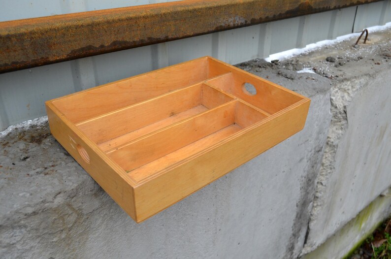 Op de afbeelding: Een lichtgekleurde houten dienblad met drie interne compartimenten en een cirkelvormige handgreepuitsparing. De lade is rechthoekig en staat op een betonnen oppervlak. Het hout heeft een natuurlijke afwerking.