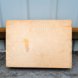 Op de afbeelding: Een rechthoekige, lichtgekleurde houten plank met zichtbare houtnerf en een kleine, onregelmatige vlek. De plank is geplaatst tegen een lichtblauwe muur en een roestige metalen balk, rustend op een betonnen oppervlak.