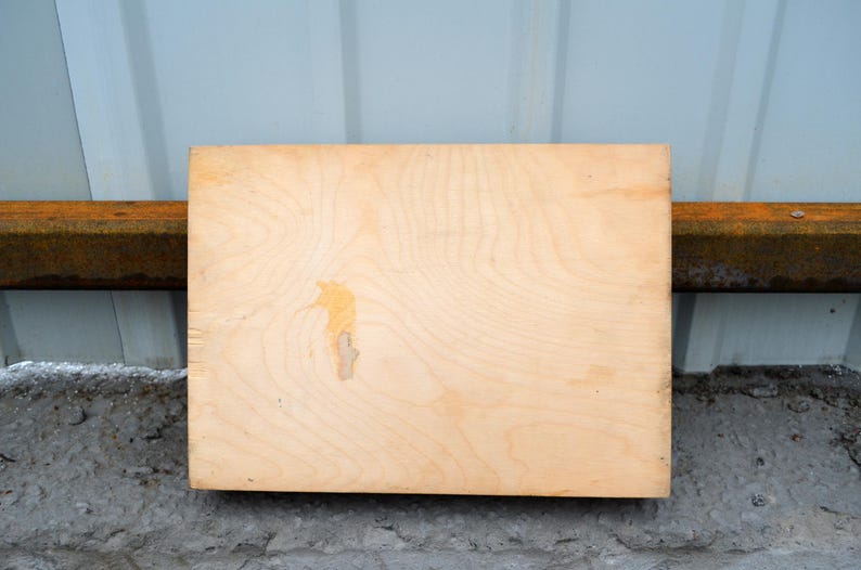 Op de afbeelding: Een rechthoekige, lichtgekleurde houten plank met zichtbare houtnerf en een kleine, onregelmatige vlek. De plank is geplaatst tegen een lichtblauwe muur en een roestige metalen balk, rustend op een betonnen oppervlak.