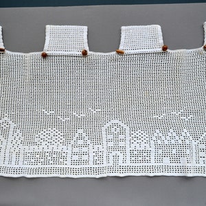 May include: A white crocheted valance featuring a grid pattern with a detailed cityscape design along the bottom edge. The top border includes four rectangular tabs, each secured with small, smooth, brown wooden beads along the top edge.