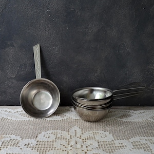 May include: A set of stainless steel measuring cups and a ladle with a decorative handle. The kitchen tools are arranged on a white lace tablecloth against a dark gray background.