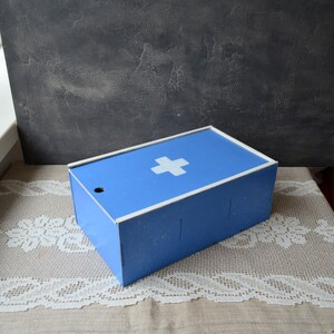 May include: A blue metal first aid box with a white cross on the lid. The box has a rectangular shape with a white trim and a small hole on the side. The box is sitting on a lace tablecloth.