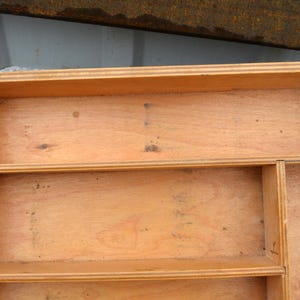 Op de afbeelding: Een lichtbruine houten opbergdoos met meerdere planken en compartimenten. De doos heeft een rechthoekige vorm met afgeronde uitsparingen aan de zijkanten. De houtnerf is zichtbaar en de doos lijkt handgemaakt.