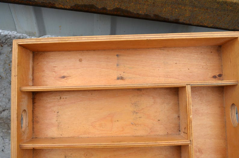 Op de afbeelding: Een lichtbruine houten opbergdoos met meerdere planken en compartimenten. De doos heeft een rechthoekige vorm met afgeronde uitsparingen aan de zijkanten. De houtnerf is zichtbaar en de doos lijkt handgemaakt.