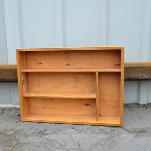 Op de afbeelding: Een houten organizer met een natuurlijke afwerking. Het heeft twee planken, een verticale verdeler en een klein handvat. De organizer is ontworpen om kleine items vast te houden en weer te geven. De houtnerf is zichtbaar.