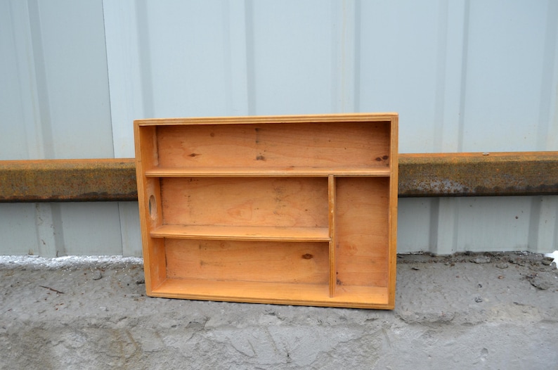 Op de afbeelding: Een houten organizer met een natuurlijke afwerking. Het heeft twee planken, een verticale verdeler en een klein handvat. De organizer is ontworpen om kleine items vast te houden en weer te geven. De houtnerf is zichtbaar.