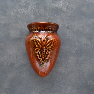 May include: A small, orange ceramic wall vase with a brown rim and a decorative embossed floral design. The vase has a glossy finish and is set against a textured grey background.