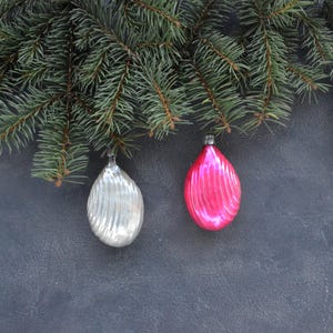 Può includere: Due decorazioni in vetro a forma di goccia appese a un ramo di pino verde. Un ornamento è argento metallizzato con texture a coste verticali, l'altro è rosa acceso con dettagli a coste verticali abbinati e un tappo in metallo argentato.