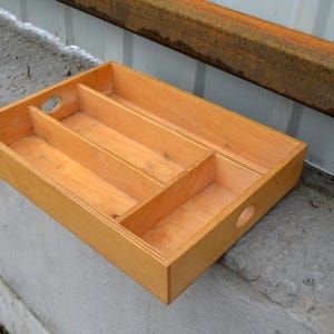 Op de afbeelding: Een rechthoekige houten bestekbak met drie compartimenten en twee ronde handgreepgaten. De bak is gemaakt van lichtgekleurd hout en is ontworpen om bestek te organiseren. De afmetingen zijn ongeveer 30 cm lang, 20 cm breed en 5 cm diep.