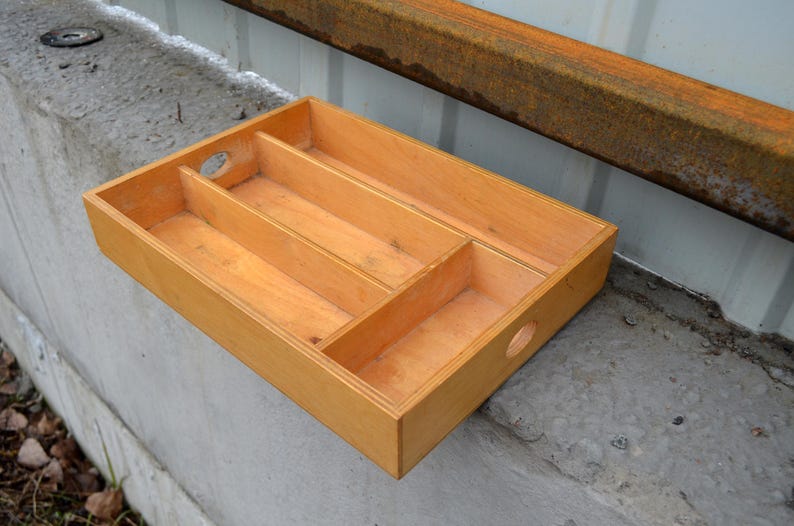 Op de afbeelding: Een rechthoekige houten bestekbak met drie compartimenten en twee ronde handgreepgaten. De bak is gemaakt van lichtgekleurd hout en is ontworpen om bestek te organiseren. De afmetingen zijn ongeveer 30 cm lang, 20 cm breed en 5 cm diep.