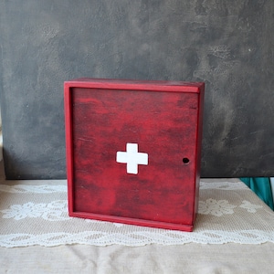 May include: A red wooden first aid box with a white cross on the front. The box has a small hole for a latch and is sitting on a cream-colored lace tablecloth. The background is a dark gray wall.