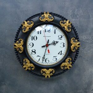 May include: A round wall clock with a black frame featuring gold-colored fleur-de-lis accents. The clock face is white with black Arabic numerals, black hour and minute hands, and a red second hand. The text "Янтарь" is printed in black.