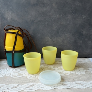 Puede incluir: Un conjunto de vasos de plástico amarillos vintage y un recipiente turquesa y amarillo con una correa de cuero marrón. Una tapa de plástico blanco se encuentra cerca de los vasos. Los artículos están sobre un mantel de encaje blanco.