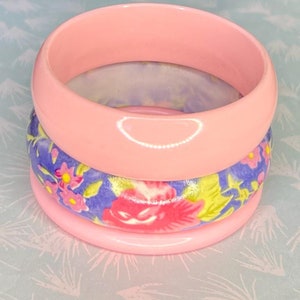 Peut inclure: Trois bracelets empilés. Les bracelets supérieur et inférieur sont rose clair uni. Le bracelet du milieu a un fond bleu avec des motifs floraux roses et jaunes. Les bracelets sont en matière lisse et brillante.