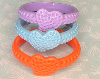 Può includere: Tre braccialetti colorati impilati uno sopra l'altro. Il braccialetto superiore è lavanda, quello centrale è azzurro e quello inferiore è arancione. Ogni braccialetto ha un accento a forma di cuore al centro e una texture a pois attorno alla fascia.