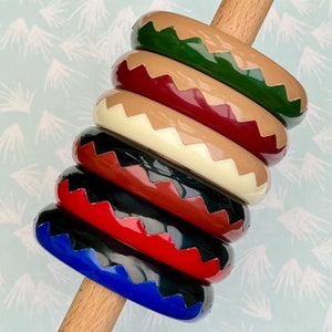 Può includere: Una pila di braccialetti colorati nelle tonalità di verde, beige, bordeaux, crema, nero, rosso e blu. Ogni braccialetto presenta un motivo a zig-zag in un colore a contrasto. I braccialetti sono esposti su un piolo di legno, mostrando i loro colori vivaci e il motivo unico.