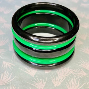 Op de afbeelding: Stapel van drie zwarte en groene armbanden. De armbanden zijn gemaakt van een glanzend materiaal, met een dikke zwarte band afgewisseld met een dunnere, felgroene band. De armbanden zijn op elkaar gestapeld en creëren een gedurfd, kleurrijk accessoire.