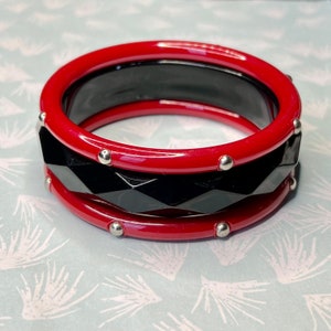 Può includere: Un set di due braccialetti rossi e neri. Le fasce superiore e inferiore sono di un rosso lucido, con piccoli borchie argentate. La fascia centrale è nera con un design geometrico sfaccettato. Gioielli.