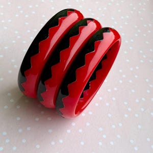 Op de afbeelding: Drie rode armbanden met een glanzende afwerking, elk met een zwart zigzagpatroon. De armbanden zijn op elkaar gestapeld, wat hun ontwerp en kleurcontrast benadrukt. Deze accessoires zijn geschikt om elke outfit op te fleuren.