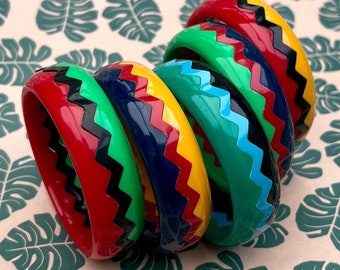 Può includere: Una collezione di braccialetti colorati con un motivo a zig-zag. I braccialetti sono rossi, verdi, gialli, blu e neri. Sono impilati l'uno sull'altro. Lo sfondo è beige chiaro con un motivo a foglie verdi.