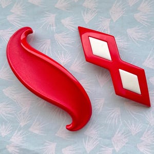 May include: Two red, decorative, vintage-style objects. One is a curved, elongated shape with a rounded end. The other is a red, double-diamond shape with white diamond inserts. Both are on a light blue background.