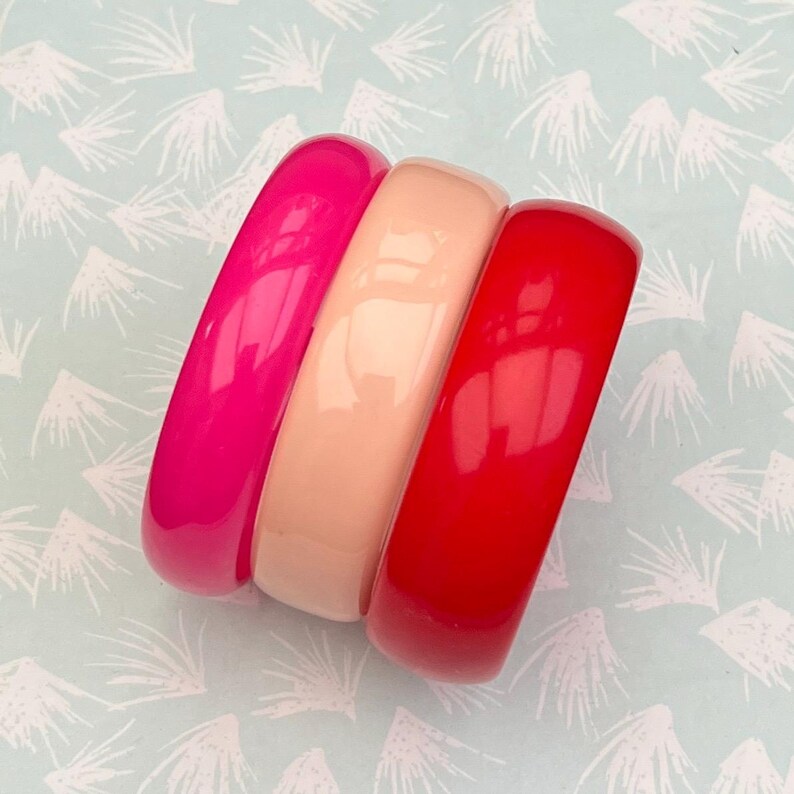 Pu&ograve; includere: Tre braccialetti lucidi nei toni del rosa, pesca e rosso. I braccialetti sono impilati, mostrando il loro design liscio e arrotondato. Lo sfondo &egrave; azzurro con un motivo bianco.
