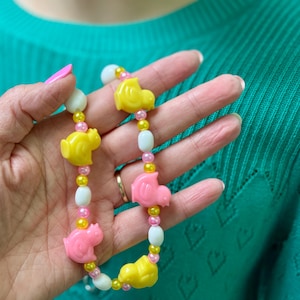 Op de afbeelding: Een ketting met gele en roze eekhoornvormige kralen, afgewisseld met witte en roze kralen. De ketting is geregen aan een fijne ketting, waardoor een speels accessoire ontstaat. De kralen hebben een gladde, glanzende afwerking.