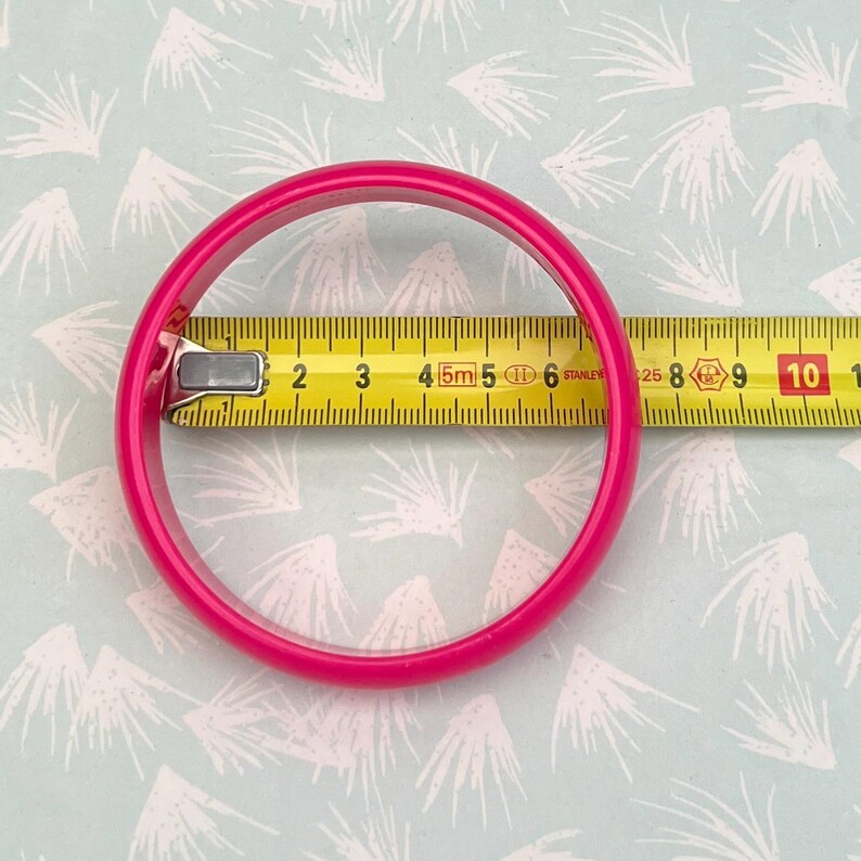 Pu&ograve; includere: Un braccialetto rosa acceso, di circa 9 cm di diametro, &egrave; mostrato accanto a un metro a nastro giallo. Il braccialetto &egrave; di colore pieno e lucido. Il metro a nastro mostra il diametro del braccialetto.