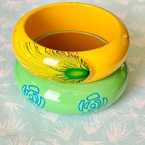 Op de afbeelding: Twee houten armbanden. De bovenste armband is geel met een pauwenveermotief in groen en geel. De onderste armband is groen met blauwe bloemmotieven. De armbanden zijn op elkaar gestapeld.