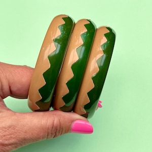 Op de afbeelding: Drie armbanden met een glanzende afwerking en een tweekleurig ontwerp. Elke armband heeft een grillig zigzagpatroon dat afwisselt tussen effen beige en diep bosgroen, wat samen een samenhangende set van cirkelvormige sieraden vormt.