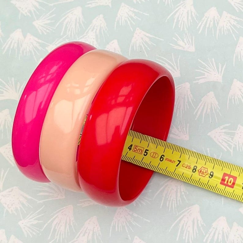 Pu&ograve; includere: Tre braccialetti lucidi rosa, pesca e rossi. Un metro a nastro giallo mostra il diametro del braccialetto rosso. I braccialetti sono un accessorio di moda. Il diametro &egrave; di circa 7,5 cm.