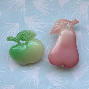 Puede incluir: Dos broches con forma de fruta: una manzana verde y una pera rosa. La manzana tiene un tallo y una hoja verdes, mientras que la pera tiene un tallo y una hoja rosas. Ambos están hechos de un material translúcido.