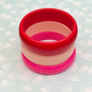 Pu&ograve; includere: Set impilato di tre braccialetti colorati. Il braccialetto superiore &egrave; rosso vibrante, seguito da un color pesca chiaro e un rosa acceso in basso. I braccialetti sono realizzati in materiale lucido e sono impilati.