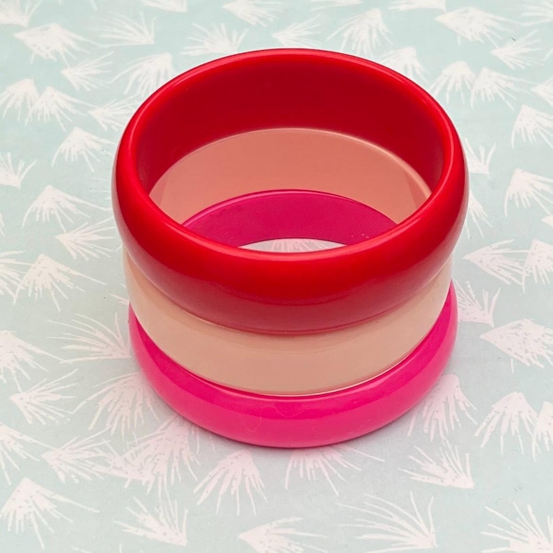 Pu&ograve; includere: Set impilato di tre braccialetti colorati. Il braccialetto superiore &egrave; rosso vibrante, seguito da un color pesca chiaro e un rosa acceso in basso. I braccialetti sono realizzati in materiale lucido e sono impilati.
