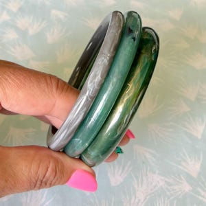 Puede incluir: Una mano sosteniendo cuatro brazaletes translúcidos en tonos de gris, verde azulado y verde. Los brazaletes tienen un diseño suave y redondeado. El fondo es un patrón azul claro y blanco.