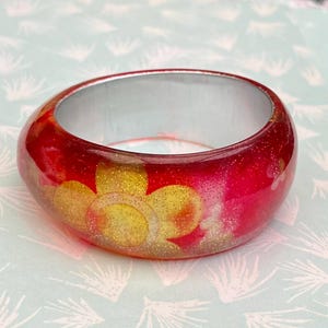 May include: A red and gold glitter bangle bracelet. The bracelet has a clear resin exterior with gold floral designs and a silver interior. The bangle is a wide, rounded shape.